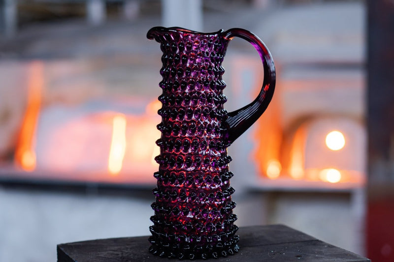 Violet Hobnail Jug Tall on a wooden base with glass ovens in the background