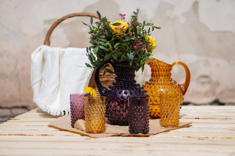 Violet Hobnail Jug on a wooden table with purple and yellow flowers, surrounded by Hobnail collection products and a wooden bag and a broken wall in the background 